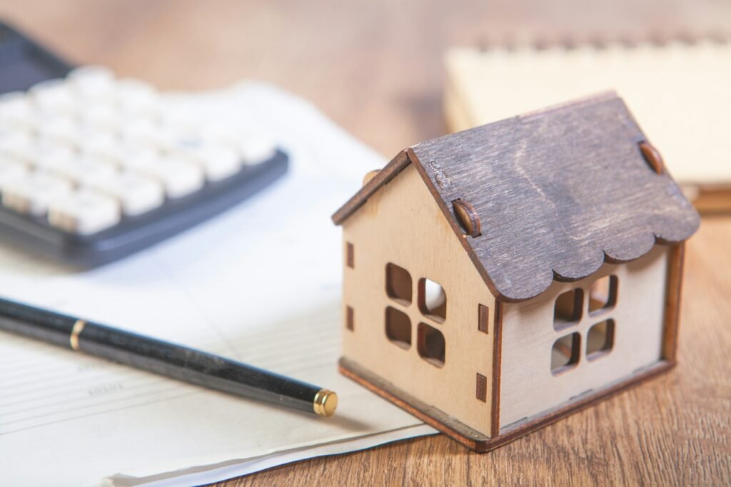 Una casita de madera junto a una calculadora, un bolígrafo y unos papeles sobre una mesa